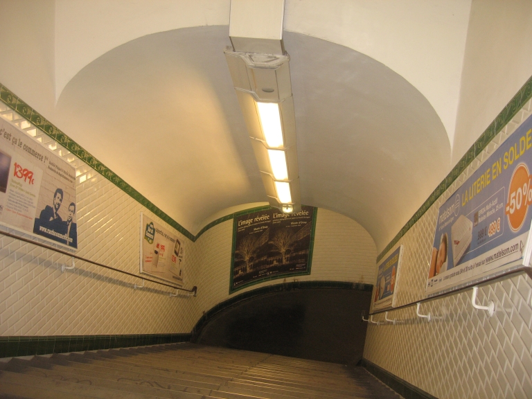 Leere gekachelte Metrotreppe, die unter einem Rundbogen in einen dunklen Gang hinabführt